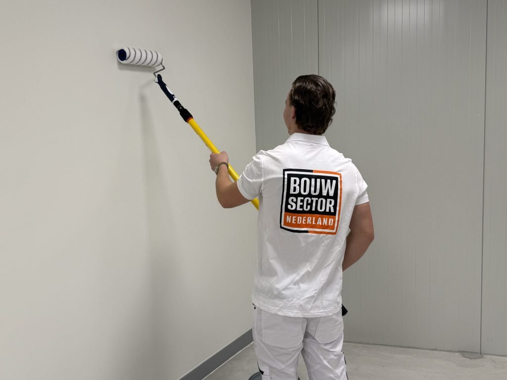 Schilder van Bouwsector Nederland begint bovenaan een lichtgrijze muur met een roller aan een lange steel