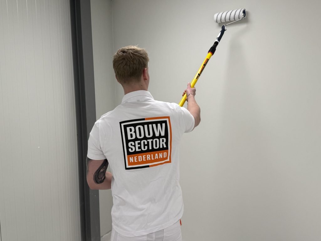 Schilder met logo van Bouwsector Nederland op zijn shirt rolt een lichte wand tot vlak onder het plafond