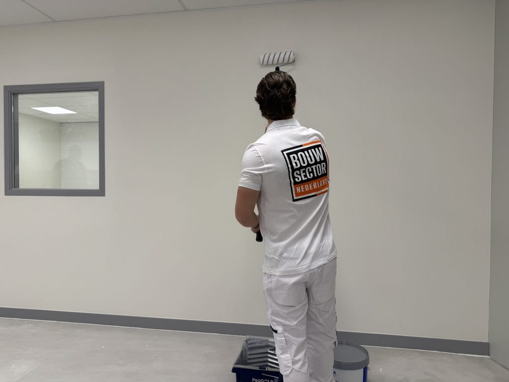 Schilder van Bouwsector Nederland zet de roller bovenaan een lichte wand, vlak onder het systeemplafond, met verfbak en emmer op de vloer