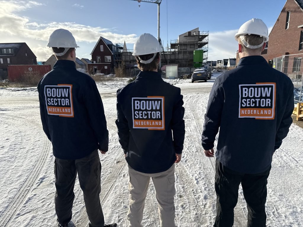 Drie vakmensen van Bouwsector Nederland met witte helm, op de rug gezien, op een besneeuwde bouwweg bij nieuwbouw in aanbouw